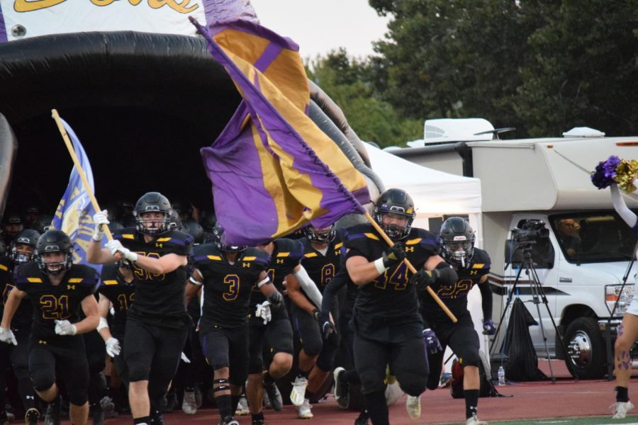 The football team runs out of the infalatable helmet, with bright flags and game day faces.