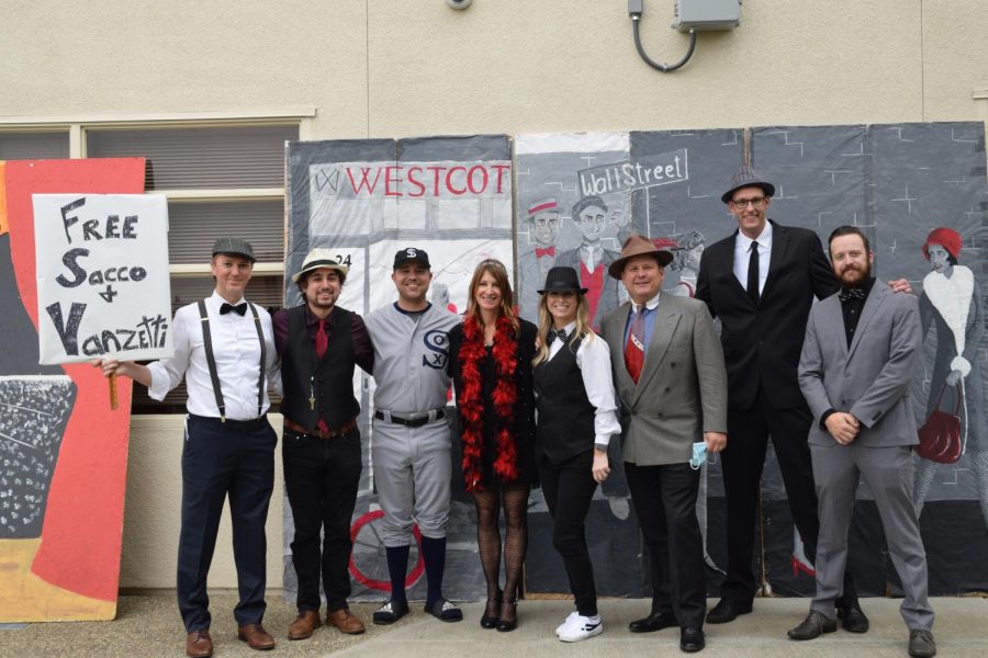 History teachers stand aside each other with big smiles, posing behind the 1920 poster and signs.