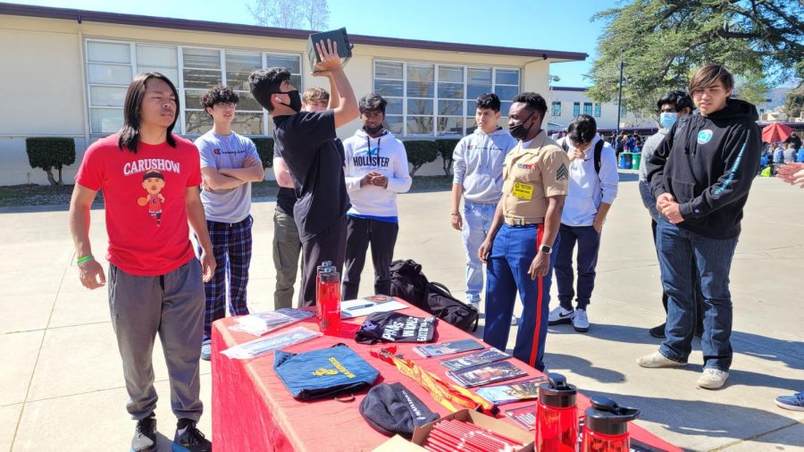 A crowd gathers as a student lifts 30 pounds of ammunition from Sergeant Mainas presentation table.