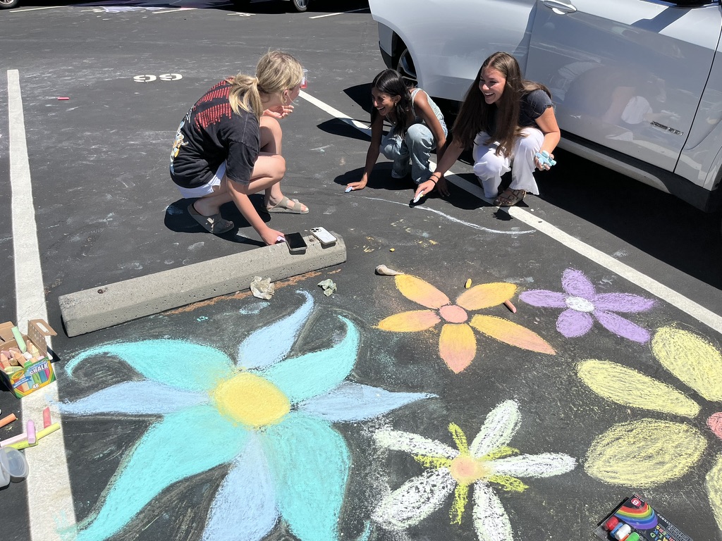 Emmy Hoopes ('26), Noor Gupta ('26), and Sarah Price ('26) talk and laugh as they draw on their parking spots.